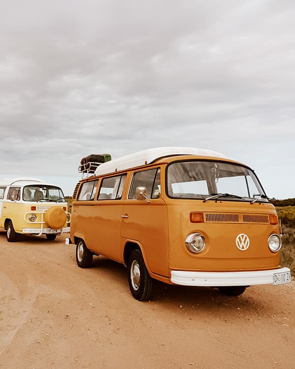 Our kombis Vera & Anna - @wanderingotooles 09/2022 Two vintage Volkswagen Kombi vans, belonging to @wanderingotooles, parked on a dusty road under an overcast sky. The orange van, featuring a roof rack loaded with supplies, is in the foreground, while the yellow van is in the background with a yellow spare tire cover. The image captures the essence of van life adventures across the Australian outback.