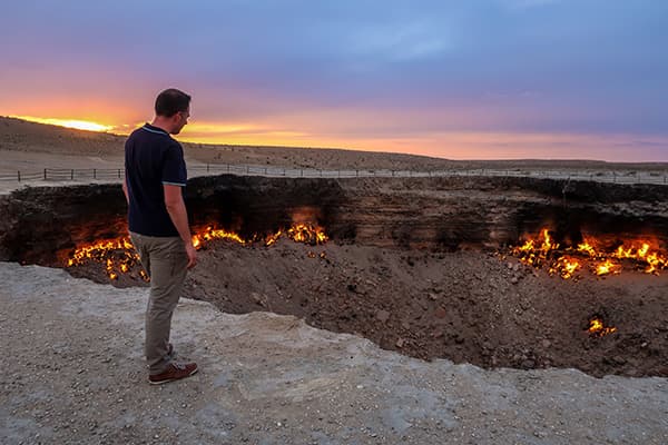At the Gates of Hell in Turkmenistan - @char.is.far 01/2025 Marcel from @char.is.far standing near the Gates of Hell, a fiery natural gas crater in Turkmenistan, during sunset. The image captures the glowing orange flames of the crater against the backdrop of a dramatic desert landscape and a colorful sky. This iconic location is a highlight of their overlanding journey through Central Asia on their route from Europe to Asia.