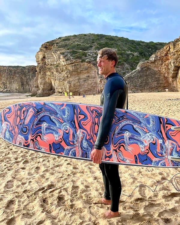 Surfing in Portugal - @van4life__ 04/2025 Chris from @van4life__ stands barefoot on a sandy beach in Portugal, holding a large, vividly patterned surfboard with swirls of pink, blue, and black. He wears a black wetsuit and smiles as he looks toward the ocean. Behind him rise rugged limestone cliffs, typical of Portugal’s Algarve coast, capturing the spirit of surf culture and vanlife.