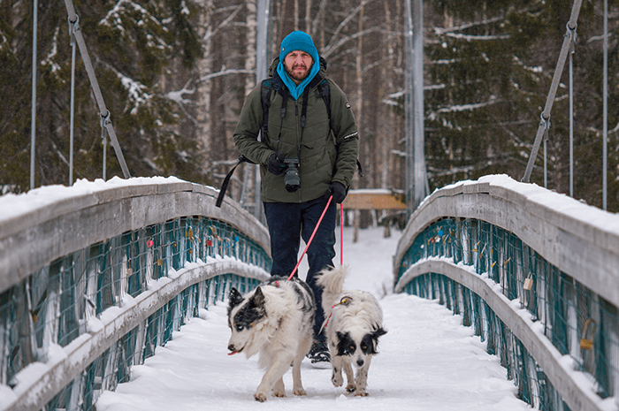A pack of three - @hrvoje_juric 12/2022 Hrvoje Juric walks with his two dogs, Ena and Max, across a snowy suspension bridge in a Finnish forest during his Dacia Duster winter vanlife adventure, holding a camera and wearing a green jacket and blue beanie, capturing the essence of his photography-focused journey through northern Europe.