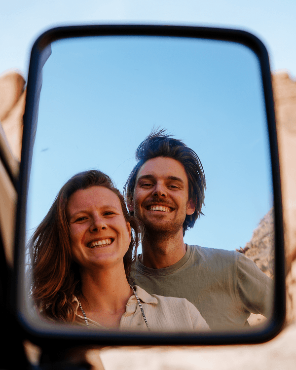 We're Amy & Jeroen - @countless_roads 03/2023 A reflection of Amy and Jeroen from @countless_roads smiling in the side mirror of their Mitsubishi L300 4x4 under a clear blue sky. The couple’s joyful expressions capture the essence of vanlife travel, freedom, and overlanding adventure during their journey across the Arabian Peninsula.