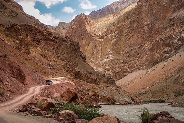 Driving the Bartang Valley in Tajikistan - @char.is.far 01/2025 @char.is.far's Toyota 4Runner navigating a narrow dirt road through the dramatic Bartang Valley in Tajikistan. The image showcases rugged red rock cliffs, a winding river, and breathtaking mountain scenery, highlighting the remote and challenging terrain of this iconic high-altitude region. This photo captures a key moment in their overlanding adventure from Europe to Asia.