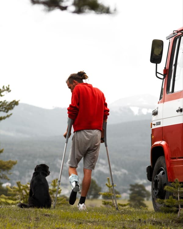 Max on crutches - @hummel_die_feuerwehr 06/2022 Max from @hummel_die_feuerwehr on crutches with a dog beside a red Mercedes-Benz 608D "Düdo" firetruck converted into a van, overlooking a scenic mountain landscape, representing the challenges of vanlife after an injury in a remote location on Crete.