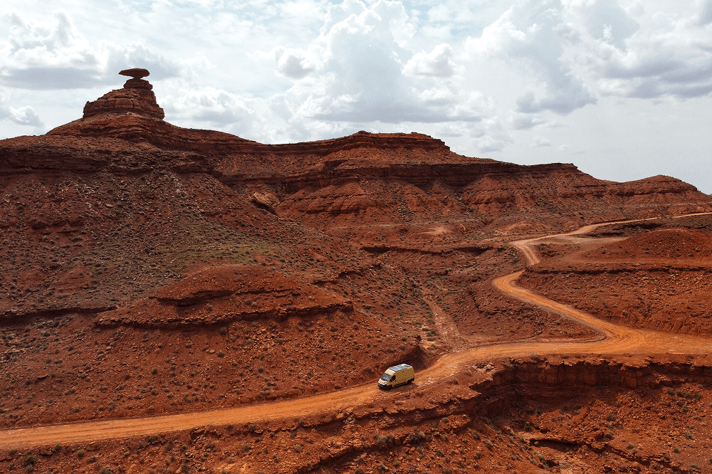 Exploring Utah - @gary_and_sheila 06/2023 Self converted Fiat Ducato camper van driving along a winding red dirt road through dramatic desert cliffs near Mexican Hat, Utah, captured during a long term vanlife and overlanding journey by @gary_and_sheila. The image shows remote canyon landscapes, rugged off road travel, and life on the road in the American Southwest under a wide cloudy sky.