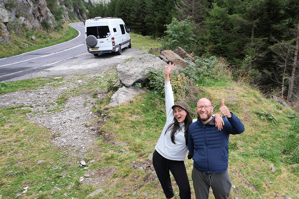 Tamara & Barry, two like minded souls - @coyama.live 03/2023 Tamara and Barry from @coyama.live smile and celebrate on a grassy mountain pull off beside their white Mercedes Sprinter camper van during their vanlife travels. They stand with arms raised in excitement near a winding alpine road surrounded by rocky cliffs and dense forest. The scene captures their joyful long term adventure and nomadic travel lifestyle.
