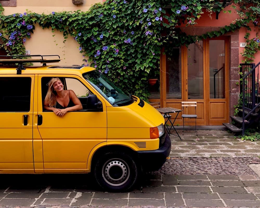 Line and Sunshine in an Italian town - @sunshinetribe_ 10/2020 A joyful woman with blonde hair leans out of the driver's side window of a bright yellow van parked on a charming cobblestone street. Behind her, a vibrant climbing plant with purple flowers adorns the exterior of a classic European building with pastel-hued walls. The scene conveys a sense of travel and adventure in a quaint urban setting, with the woman's smile suggesting a carefree and exploratory spirit.