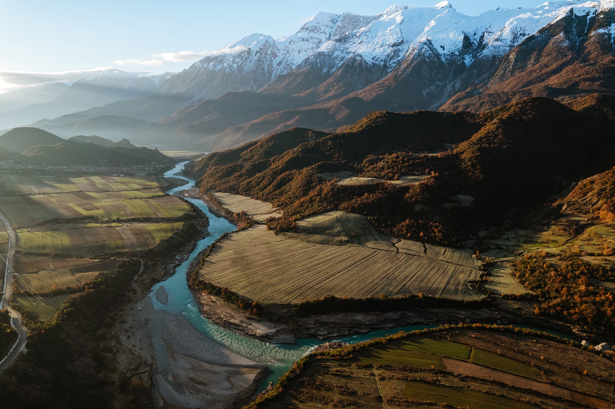 Aerial view of Permet, Albania - @edelweiss.on.the.road 06/2022 Aerial view of Permet, Albania, with the Vjosa River winding through vast agricultural fields and lush, rolling hills. Snow-capped mountains rise majestically in the background, under a clear sky with soft sunlight casting long shadows over the diverse landscape. This drone shot captures Permet’s natural beauty, highlighting Albania’s unspoiled countryside, with the Vjosa River as a focal point, showcasing the region’s rugged charm and rich geographic diversity in the Balkans.
