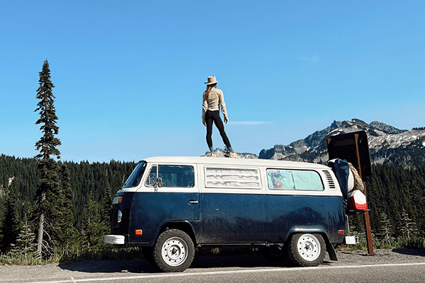 A monumental commitment - @wanderingnwonder 06/2025 Tara of @wanderingnwonder stands confidently on the roof of their navy and white 1978 Volkswagen Type 2 Kombi, parked on a scenic mountain road surrounded by evergreen trees and rugged peaks. This image captures the spirit of freedom, adventure, and vanlife exploration through the wild landscapes of the USA.