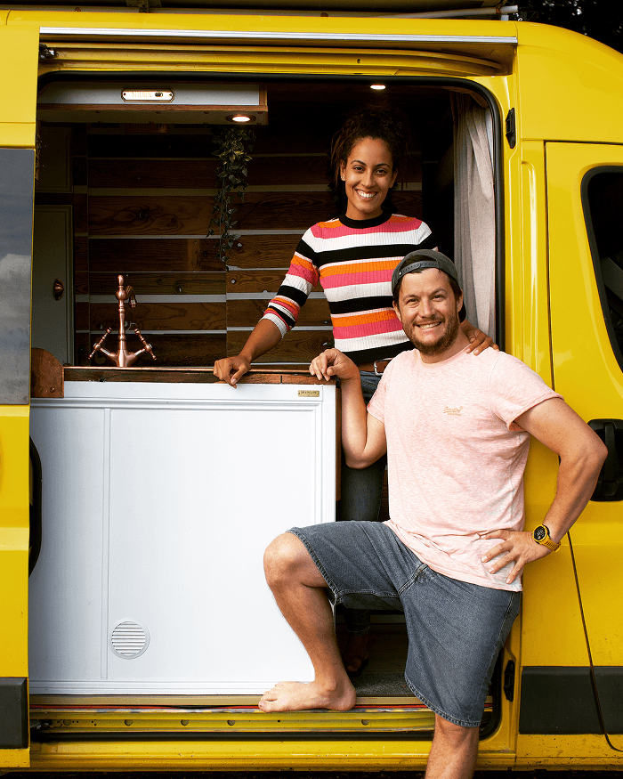 Gary & Sheila in their van - @gary_and_sheila 06/2023 Gary and Sheila, the vanlife couple behind @gary_and_sheila, standing barefoot in the open sliding door of their bright yellow self converted Fiat Ducato camper van. The image shows the custom van interior with wooden panels, compact kitchen, copper faucet, and cozy details, highlighting full time vanlife, DIY van conversion, and life on the road during their long term overland journey.