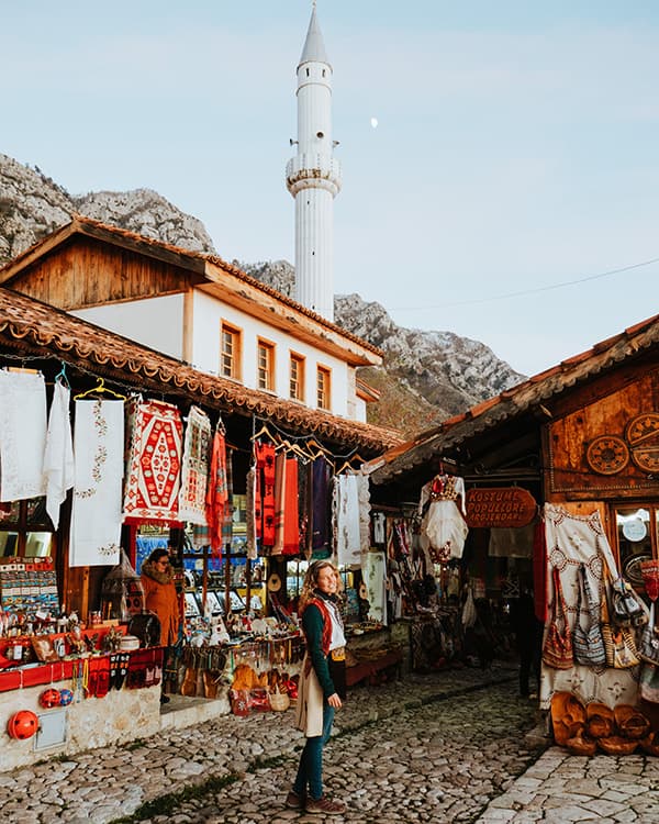 A traditional bazaar in Kruja, Albania - @edelweiss.on.the.road 06/2022 Regina from @edelweiss.on.the.road exploring a traditional bazaar in Kruja, Albania. The image captures colorful handmade textiles, souvenirs, and traditional crafts displayed in wooden market stalls against a backdrop of historic Ottoman architecture and a tall minaret. The mountainous landscape of Kruja frames this vibrant Albanian marketplace, showcasing the cultural richness and artisanal heritage of the town.