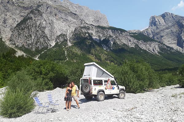 A journey to places where solutions are already growing - @advandortmontduurzaam 04/2025 Ad van Dortmont’s white Land Rover Defender 110 overland camper parked on a rocky riverbed surrounded by lush green forest and dramatic limestone mountains in the Balkans. The vehicle's pop-up roof is extended, and the side features expedition stickers. Two people stand smiling in front of the Land Rover, with two blue camping chairs set up nearby, under clear blue skies. The scene captures the spirit of vanlife, overlanding, and sustainable travel through remote and wild landscapes.