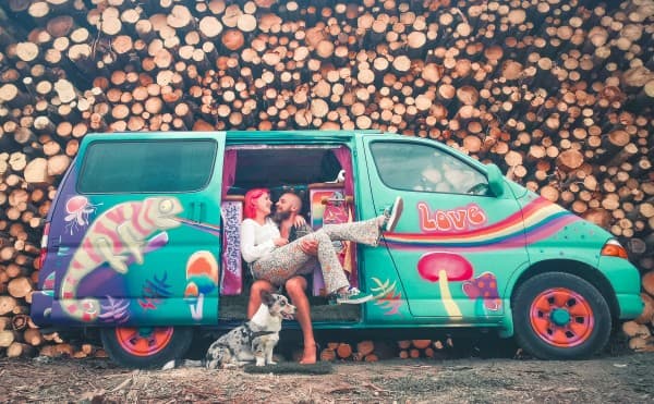 Sandra, Joriën & Fudge with their Toyota camper van - @travelwildtobefree 08/2021 Joriën and Sandra from travelwildtobefree, sitting happily in their colorful, hippie-themed Toyota HiAce 4x4 camper van with their corgi Fudge, in front of a stacked pile of logs.