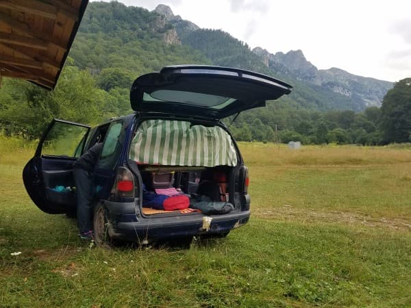 Camping in the Renault Espace - @feel_free_van 02/2022 A Renault Espace microcamper parked in a lush green meadow, surrounded by mountains in Bulgaria. The back of the van is open, revealing a cozy, makeshift sleeping area with bedding and supplies. A person is seen arranging items inside the vehicle, preparing for a peaceful night in nature. The scene captures the essence of a simple and adventurous van life in a mountainous region.