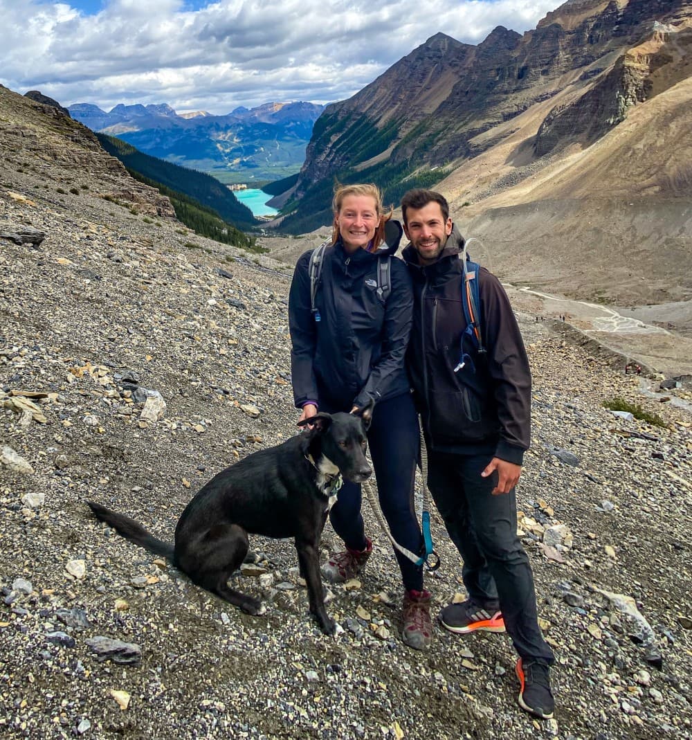 The trio on a hike at Lake Louise - @hayleyandjake 10/2020 A smiling couple with a black dog standing on a rocky trail at Lake Louise, with the vibrant turquoise lake visible in the valley below, surrounded by majestic mountain ranges and a dramatic sky with patches of blue and cloud cover.