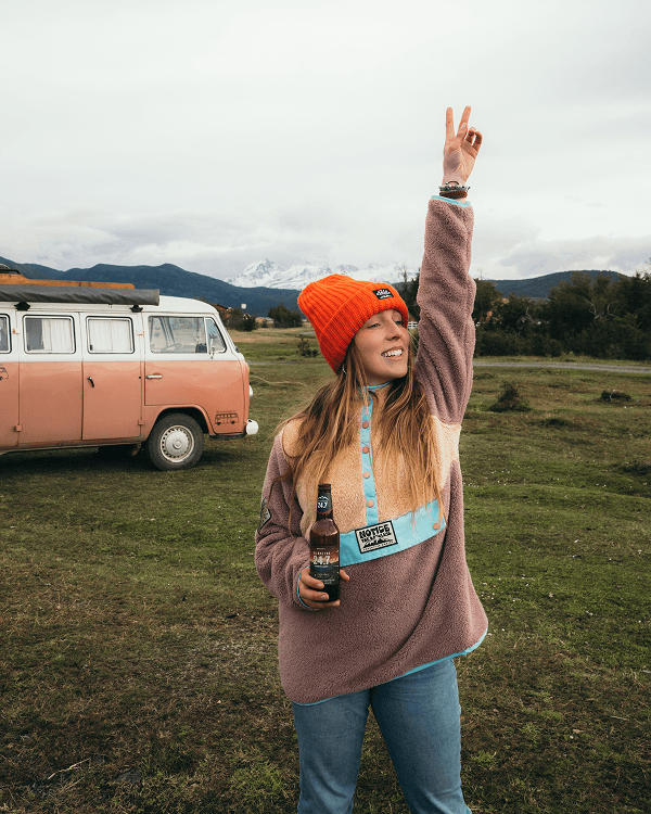 I realized I should try it alone - @octaviaviajando 09/2022 Victoria from Chile, known as @octaviaviajando on Instagram, smiles while holding a craft beer and raising a peace sign in an open grassy field near Patagonia. She wears a bright orange beanie and a cozy fleece pullover, standing next to her iconic pink and white 1985 Volkswagen T2 Kombi campervan named Octavia. Snow-capped peaks rise in the distance under a cloudy sky. This moment captures her joyful vanlife spirit, slow travel lifestyle, and connection to nature and adventure.