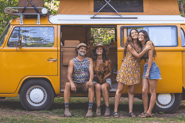 Adventures together in our vintage kombi - @summerofseventyfive 12/2022 The summerofseventyfive family sits and stands beside their bright yellow 1975 Volkswagen Kombi camper van with its pop-top roof raised, parked in a lush Australian forest. The parents sit in the open side doorway with their dog, while their two daughters stand nearby smiling and hugging. The roof rack is loaded with vintage suitcases and gear, reflecting a warm and joyful vanlife lifestyle filled with family travel, adventure, and connection to nature.