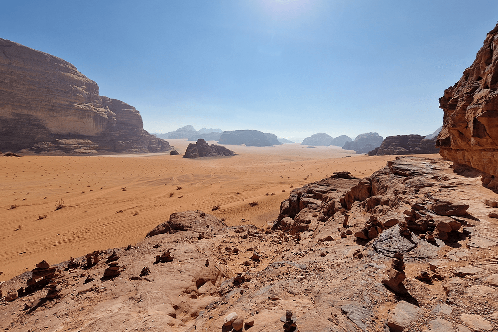 The Wadi Rum desert in Jordan - @paul.jaunegg 03/2023 Expansive view of the Wadi Rum desert in Jordan with reddish sand dunes, rugged sandstone cliffs, and distant rock formations under a bright blue sky. The scene captures the vastness and beauty of this iconic Middle Eastern landscape. Photo by Paul Jaunegg.