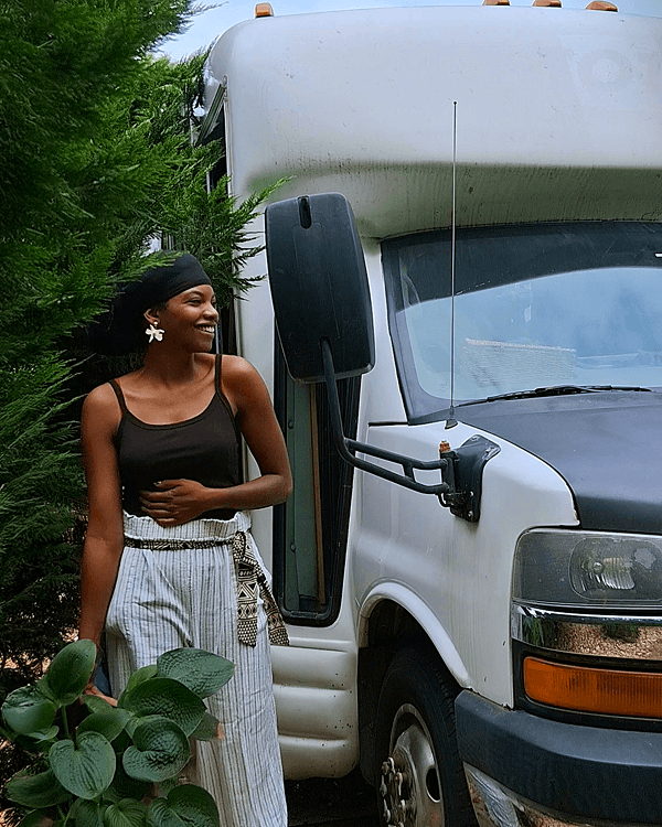 Allison Leah & her Chevrolet E3500 "Lupine" - driftwoodartwork 06/2025 Allison Leah Thompson stands beside her white Chevrolet E3500 shuttle bus "Lupine". Wearing striped pants and a dark tank top, she appears relaxed outside her mobile art studio and home. The image reflects her life as a solo female traveler and nomadic charcoal artist. Instagram handle @idonthavegum.