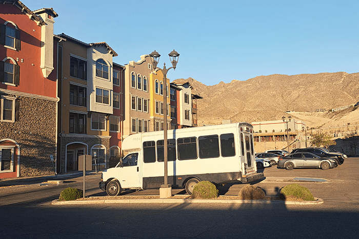 Lupine in El Paso, Texas - driftwoodartwork 06/2025 The white Chevrolet E3500 shuttle bus named Lupine is parked in a sunlit lot in El Paso, Texas, beside a row of colorful apartment buildings with desert mountains in the background. The bus belongs to charcoal artist Allison Leah Thompson, known on Instagram as @idonthavegum, who travels the USA in this converted mobile art studio.