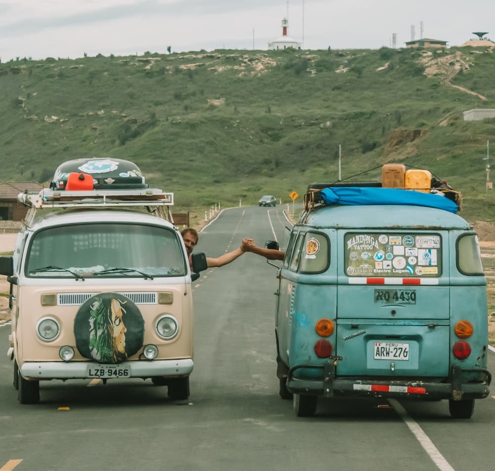 New friends & new experiences - @enjoytripbr 10/2020 Two vintage Volkswagen vans parked on a paved road, with their drivers reaching out for a handshake between the vehicles. One van is cream-colored with a large artistic face painting on the spare tire cover, while the other is blue with a collection of travel stickers and a surfboard on the roof. In the background, there's a hilly landscape with a lighthouse, under an overcast sky, suggesting a spirit of camaraderie and travel adventure.