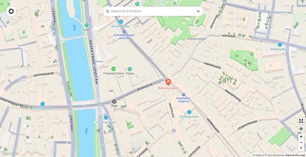 Enterprise locations stand out even more on vanlifezone.com/map Screenshot of an enterprise business location displayed on the map view of vanlifezone.com. The map shows a detailed urban area alongside a river running through the city. Various points of interest are marked, such as drinking water stations and charging stations for travelers. The specific business location is highlighted with an orange pin labeled 'Demo Location,' surrounded by dense building layouts and nearby landmarks.