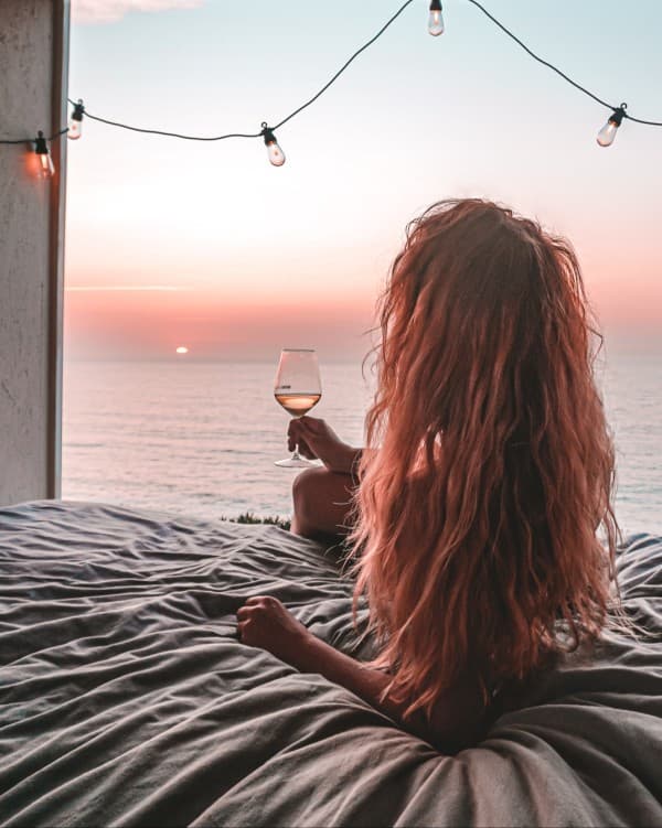 Enjoying the sunsets in Greece - @offthemainroad_ 08/2021 A blond woman enjoying a sunset view over the ocean from the back of a converted box truck camper, with string lights and a glass of wine.