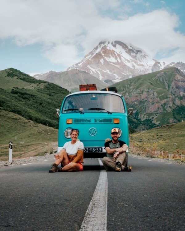 Milene & Yuri with their VW T2 "Alexine" - @mygrations.nl 02/2022 A man and a woman, Milene and Yuri from @mygrations.nl sitting on the road in front of a turquoise Volkswagen T2 van with a mountainous landscape in the background. The van is parked on a clear road with the scenic snow-capped peaks of the Greater Caucasus Mountains, possibly near Mazeri, Georgia, in the distance. The bright blue sky with scattered clouds enhances the adventurous spirit of their journey along the ancient Silk Road. The image captures the essence of van life and road trips through remote and beautiful landscapes.
