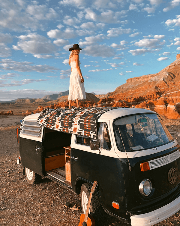 Tara quickly became a savvy camp spot scout - @wanderingnwonder 06/2025 Tara of @wanderingnwonder stands barefoot on the roof of their black and white 1978 Volkswagen Type 2 Kombi, gazing out over red rock formations and desert landscapes under a sky filled with soft clouds. A patterned blanket is spread across the van roof, and a guitar rests on the ground nearby, capturing the spirit of freedom, creativity, and vanlife in the American Southwest.