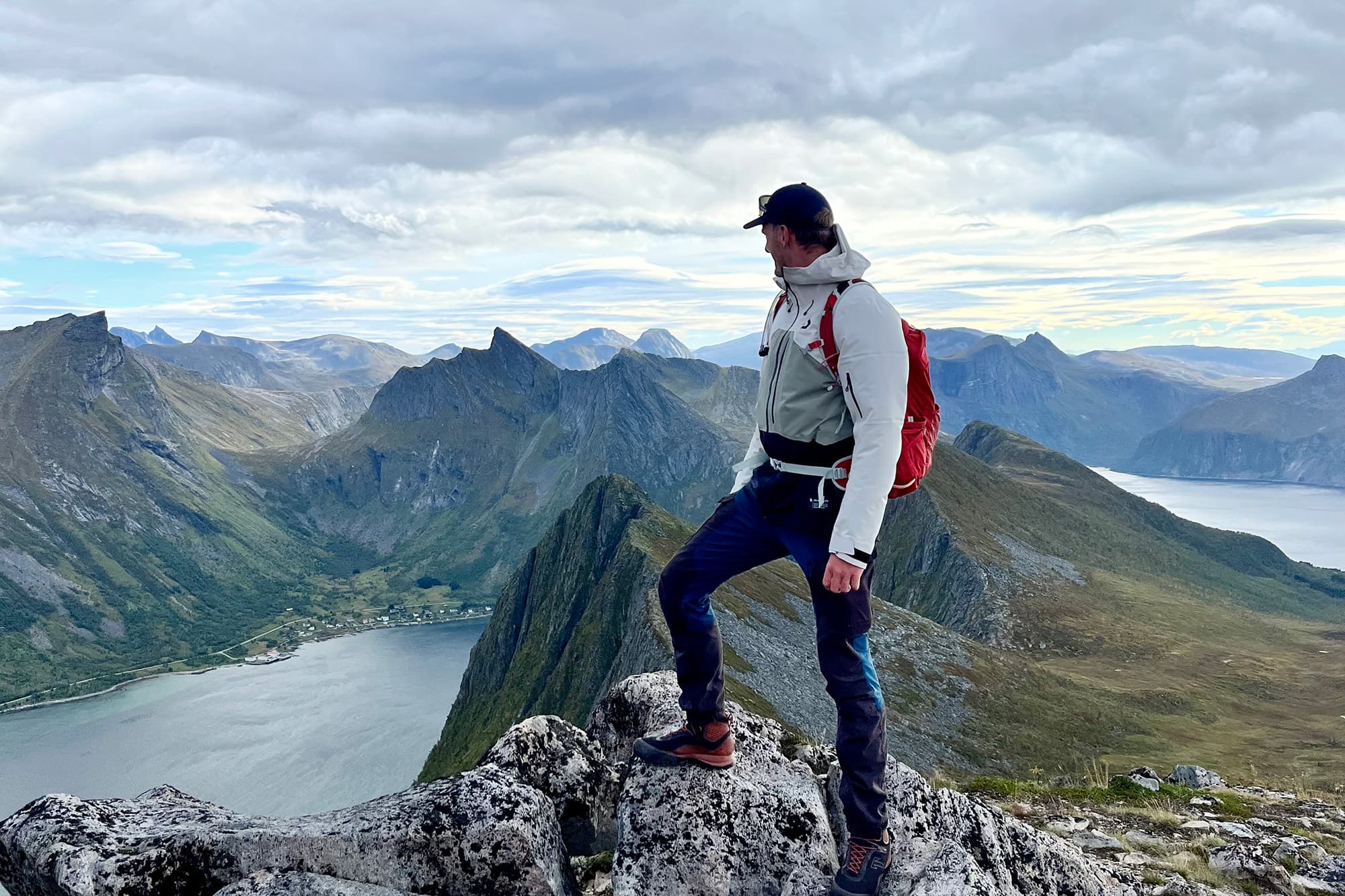 Chris exploring Norway by himself - @van4life__ 10/2024 Chris (@van4life__) standing on a rocky peak during a hike in Norway, overlooking stunning mountain ranges and a fjord. The image captures the expansive natural beauty of Norway's dramatic landscapes, with Chris in outdoor gear and a red backpack, embodying the spirit of adventure and exploration.