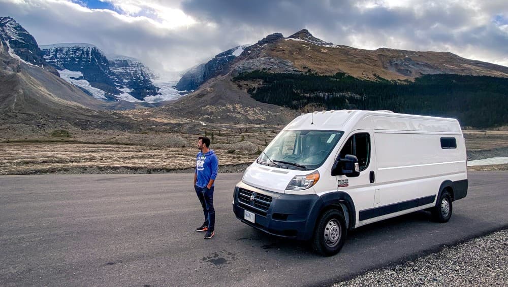 Exploring the Icefields Parkway with Rambo - @hayleyandjake 10/2020 A man standing by a Ram Promaster 159 Extended van parked on the Icefield Parkway with dramatic overcast skies above and rugged mountain scenery in the background, conveying the majestic and wild nature of the Canadian Rockies.