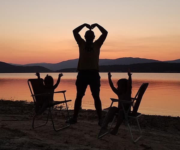 Family sunset by the lake - @feel_free_van 02/2022 A silhouette of a man and two young girls enjoying the sunset by a serene lake during their van life adventure in Bulgaria. The man stands in the middle, forming a heart shape with his hands above his head, while the girls sit in camping chairs on either side, raising their arms joyfully. The scene captures a peaceful and loving moment at the end of a day spent in nature.