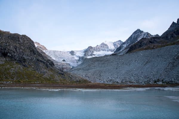 Valais, Switzerland - @miles_together 08/2021 A stunning view of a glacier in the Valais region of Switzerland, with rocky mountains and snow-capped peaks in the background. In the foreground, a serene glacial lake reflects the surrounding landscape under a clear sky. This image captures the natural beauty and rugged terrain of the Swiss Alps, showcasing the breathtaking scenery that Christina and Ralf from @miles_together explore during their adventures.