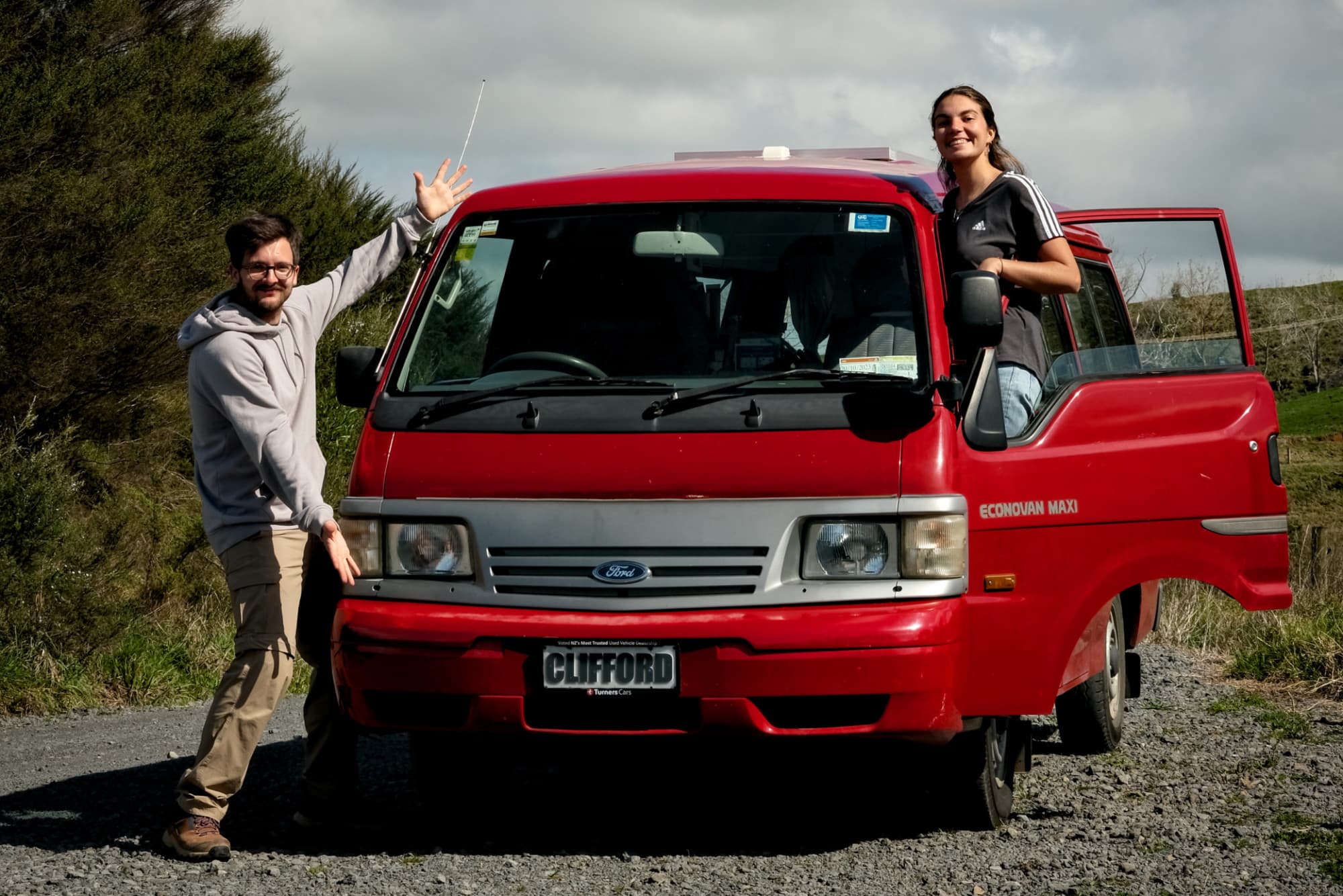 David, Claire and their van Clifford in New Zealand - @carnet_declaireurs 06/2024 David and Claire posing with their red Ford Econovan named Clifford, parked on a gravel path amidst lush greenery in New Zealand. David gestures enthusiastically towards the van while Claire leans out of the passenger door, showcasing their adventurous vanlife journey. This image captures the spirit of exploration and travel photography by the authors, @carnet_declaireurs.