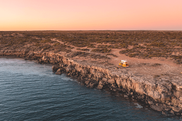 Coastal sunset from our vintage kombi - @summerofseventyfive 12/2022 The summerofseventyfive family parks their bright yellow 1975 Volkswagen Kombi camper van on a rugged cliff edge overlooking the deep blue waters of the Australian coastline during a pink and orange sunset. The pop-top roof is raised and the family sits outside enjoying the view, surrounded by rocky terrain and untouched coastal wilderness. This serene vanlife scene captures the essence of remote travel, family adventure, and the beauty of Australia’s wild shores.
