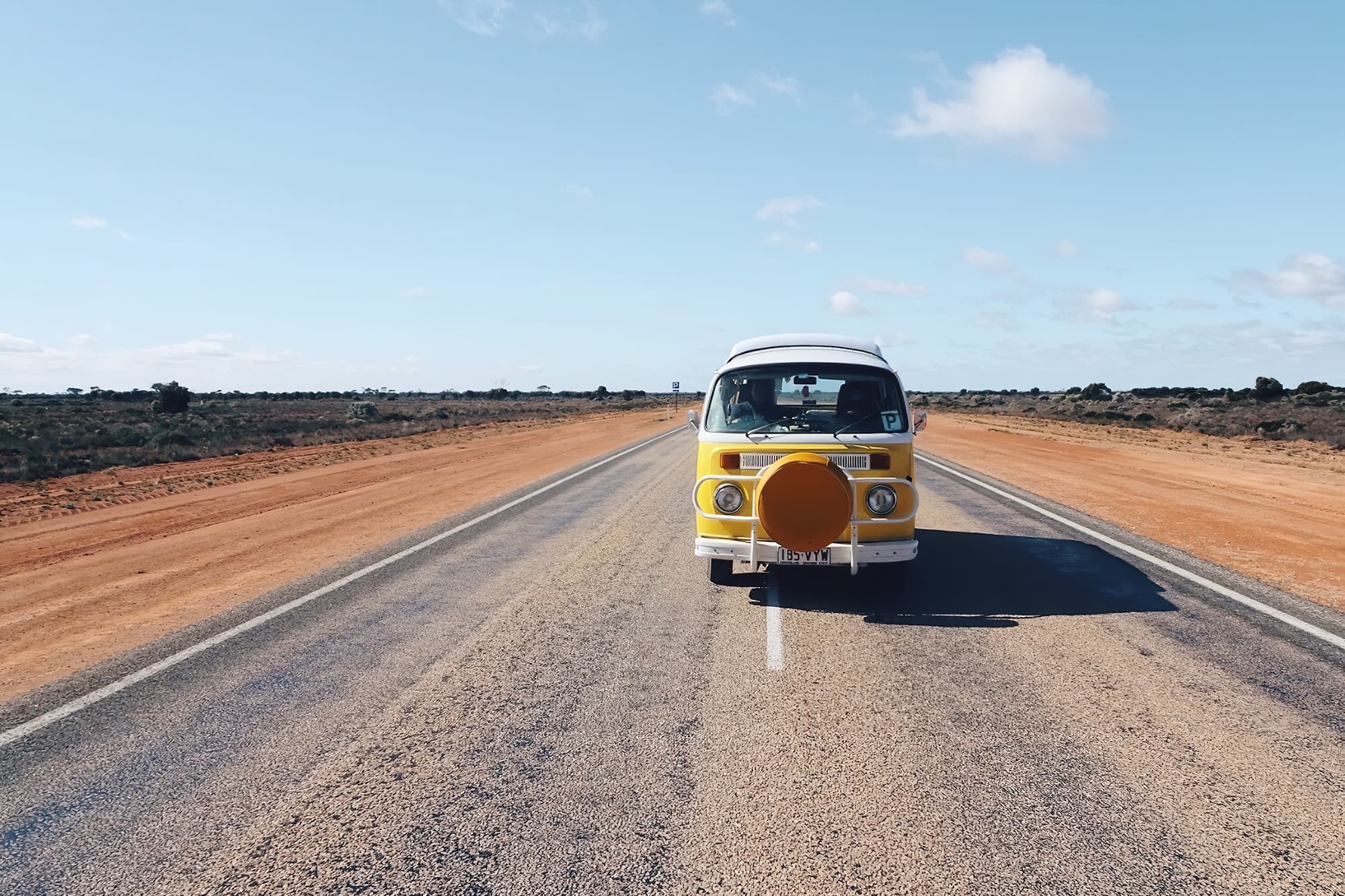 Anna on the open road - @wanderingotooles 09/2022 A vintage yellow Volkswagen Kombi van, belonging to @wanderingotooles, parked in the center of a straight, empty road in the Australian outback under a bright blue sky. The van features a yellow spare tire cover, with red dirt and sparse bushland lining both sides of the road.