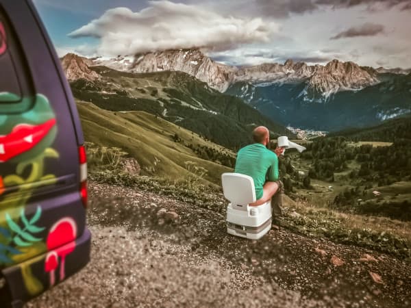 Taking in the views, one roll at a time - @travelwildtobefree 08/2021 Joriën from travelwildtobefree, sitting on a portable toilet with a roll of toilet paper, enjoying a scenic mountain view from their colorful camper van.