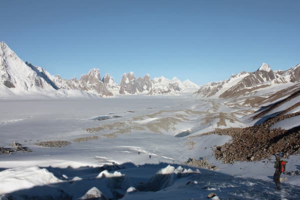 The Karakoram Mountains of Pakistan - @advandortmontduurzaam 04/2025 Wide view of the Biafo Glacier in the Karakoram Mountains of Pakistan, showing a vast, frozen river of ice stretching into the distance, framed by rugged, snow-covered peaks under a clear blue sky. Jagged mountain ridges and sharp nunataks (rocky peaks protruding through the glacier ice) rise dramatically from the icy landscape. A lone trekker with a backpack stands on the rocky moraine at the glacier’s edge, emphasizing the scale and isolation of the environment. This high-altitude glacier region is part of the third-largest ice reserve outside the polar areas and plays a key role in local water storage and climate adaptation efforts, including manmade icetower projects.