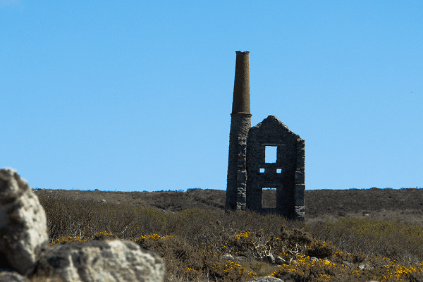 Ruins of an old Cornish tin mine engine house - @joshjmcclean 12/2022 Ruins of an old Cornish tin mine engine house with a tall brick chimney standing on a windswept heath under a bright blue sky, surrounded by yellow gorse bushes, symbolizing Cornwall’s rich mining history and heritage.