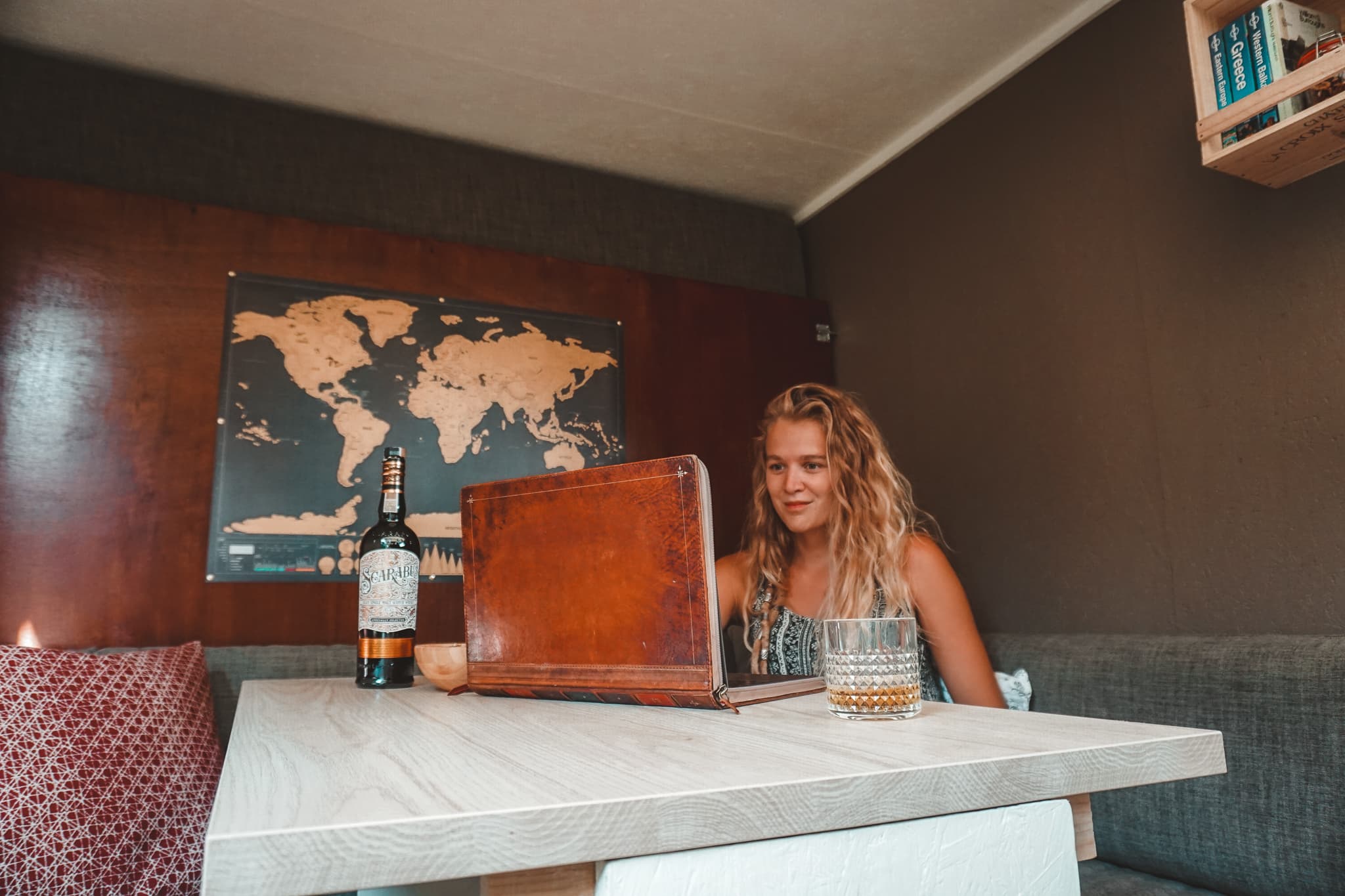 Charlotte working inside of their van - @offthemainroad_ 08/2021 Young woman working on a laptop with a vintage book-style cover, seated at a table inside a tiny home. The table features a bottle of Scotch and a glass of whiskey. In the background, a world map hangs on the wall, adding a cozy, traveler's vibe to the compact, wood-paneled interior.