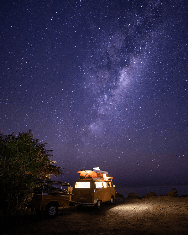 We wanted something different - @summerofseventyfive 12/2022 The summerofseventyfive family’s bright yellow 1975 Volkswagen Kombi camper van is parked beside a palm tree on the Australian coast under a breathtaking night sky filled with countless stars and the glowing band of the Milky Way. The van’s pop-top roof is open and warm interior lights glow softly, casting light onto the sandy ground. A small trailer is attached, and the ocean lies in the background, creating a magical vanlife scene of off-grid camping, stargazing, and the beauty of Australia’s wilderness.