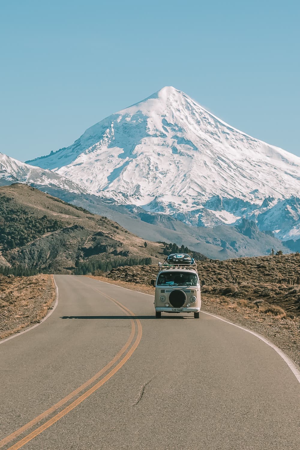 The road teaches us a lot every day - @enjoytripbr 10/2020 A classic Volkswagen van with luggage on the roof rack drives on a scenic highway away from a majestic, snow-capped mountain. The road curves gently in the foreground, framed by rugged terrain and clear blue skies, invoking a sense of adventure and travel in the great outdoors.