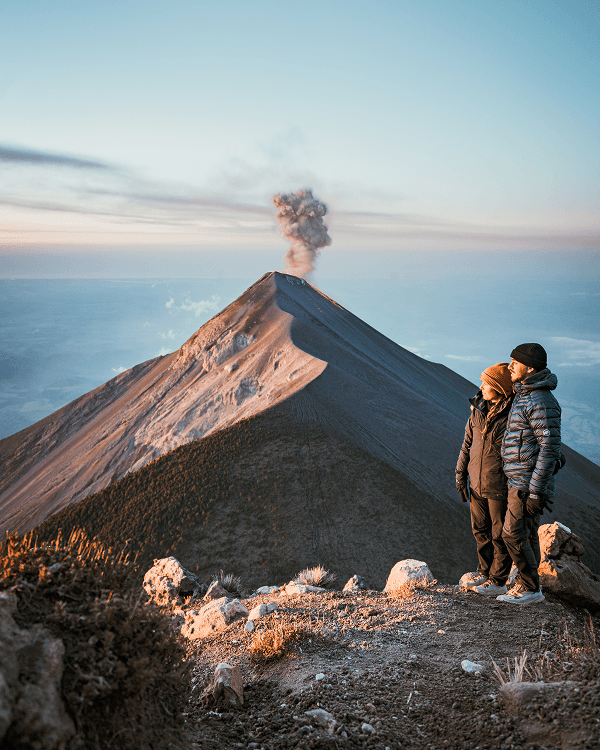 Watching Guatemala’s active Volcán de Fuego erupt - @on.vagabonde 03/2023 Audrey and Benji from on.vagabonde stand on a mountain ridge at sunrise watching Guatemala’s active Volcán de Fuego erupt with smoke, during their Pan American Highway vanlife journey.