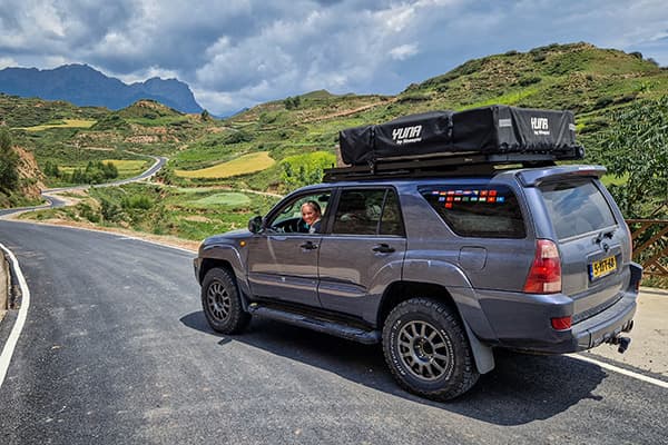 Driving through China - @char.is.far 01/2025 Charelle from @char.is.far driving their Toyota 4Runner with a rooftop tent on a winding mountain road in China. The lush green hills, terraced fields, and dramatic mountain backdrop showcase the scenic landscapes encountered during their overlanding journey from Europe to Asia. The vehicle is adorned with country flag stickers, symbolizing the extensive travel adventure.
