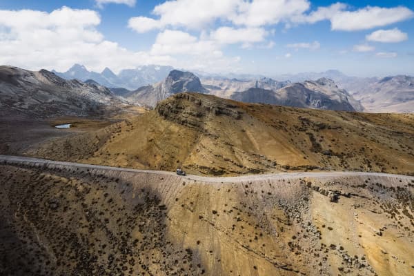 Driving through the majestic peaks of the Andes - @_roadtales 11/2024 A rugged Puch 230GE camper driving along a narrow mountain road through remote, rocky terrain in the Andes, South America, with expansive mountainous landscapes in the background.
