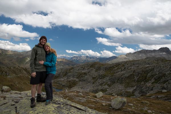 Christina & Ralf in Gotthard, Switzerland - @miles_together 08/2021 Christina and Ralf, known as @miles_together, are standing together and smiling on a rocky outcrop in the Gotthard region of Switzerland. They are surrounded by a stunning mountainous landscape under a partly cloudy sky. The image captures their adventurous spirit and love for exploring nature.