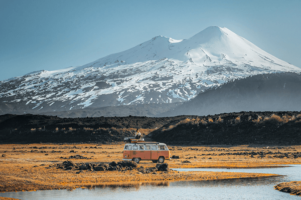 I began to relate to tourism - @octaviaviajando 09/2022 Victoria, known as @octaviaviajando, stands on top of her vintage pink and white 1985 Volkswagen T2 Kombi campervan named Octavia. The van is parked beside a clear stream in a golden high-altitude field with a dramatic snow-covered volcano rising in the background under a blue sky. Captured in the Chilean Andes, this moment highlights her solo journey through remote landscapes, celebrating slow travel, adventure, and a deep connection with nature.