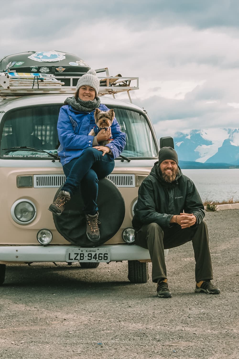 Roana, Diego & Zed - @enjoytripbr 10/2020 A smiling woman wearing a blue jacket and a beanie sits on the front tire of a vintage cream-colored Volkswagen van, holding a small Yorkshire Terrier. A man with a beard, also smiling, sits on the ground leaning against the van, wearing a black beanie and jacket. They seem to be enjoying a chilly day with a scenic backdrop of mountains and a cloudy sky. The van has a roof rack with assorted gear, suggesting an adventurous road trip.