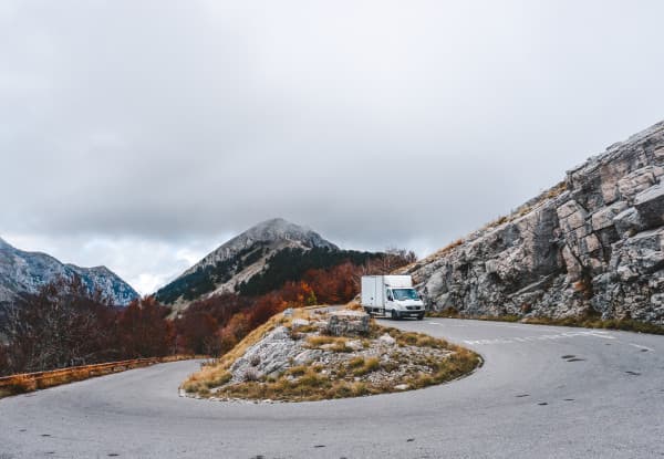 Exploring new territories - @offthemainroad_ 08/2021 White box truck driving on a winding mountain road with rocky terrain and autumn-colored trees, under a cloudy sky. The road curves around a hill with a prominent peak in the background, showcasing a dramatic and scenic mountainous landscape.