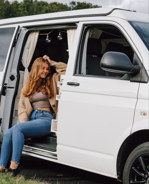 Enjoying the van - @itsme.jenni 02/2022 A woman (@itsme.jenni) sitting in the doorway of her self-built camper van, enjoying the outdoors. She is smiling and relaxed, with the van's interior partially visible behind her.
