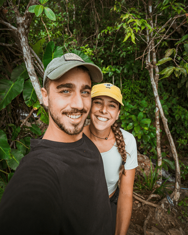 We're Audrey & Benji - @on.vagabonde 03/2023 Audrey and Benji from on.vagabonde smile while taking a selfie in a lush green jungle during their Pan American Highway vanlife journey, wearing casual outdoor clothing and caps.
